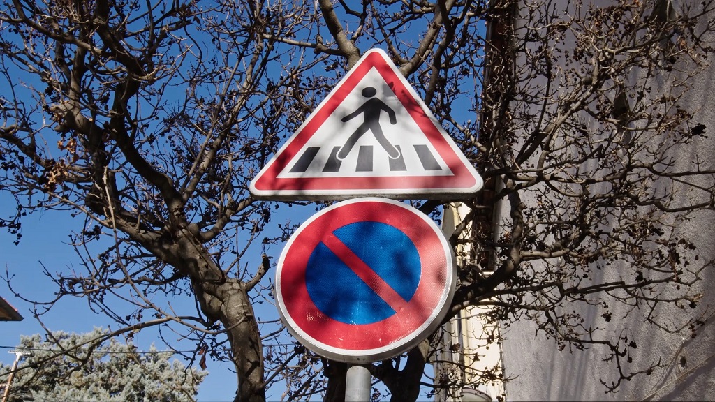 Znak pešačkog prelaza iznad znaka zabrane parkiranja ispred drveća bez lišća